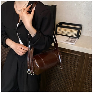 Detail of glossy dark coffee cylindrical shoulder bag worn by model against neutral wall