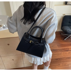 Matte black structured handbag with gold hardware shown on a shoulder in a minimalist setting