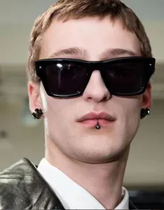 Close-up of black square sunglasses with silver rivets on a male model with ear and lip rings