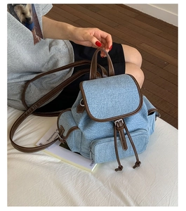 Light blue denim backpack with brown leather trim and drawstring flap closure on white bed