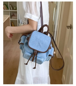 Detail of light blue denim backpack with brown leather trim worn by a model in a room