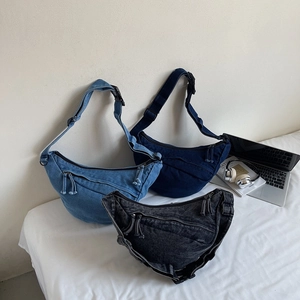 Three colors of denim crescent crossbody bags displayed with lifestyle accessories on a bed