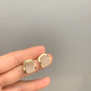 Dettaglio di orecchini a perno quadrati dorati con inserto glitter rosa su sfondo grigio
