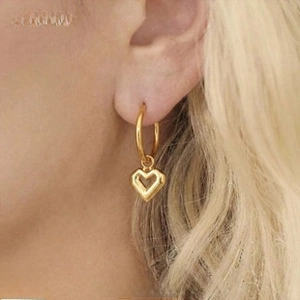 Detail of a woman wearing gold-plated C-shaped earrings with a hollow heart charm pendant