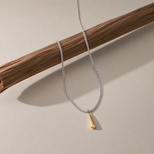 Collana minimalista bicolore con ciondolo a goccia esposta su un ramo di legno testurizzato