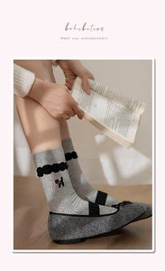 Close-up of grey crew socks with cat embroidery, lace trim, and bow shoes in a cozy setting