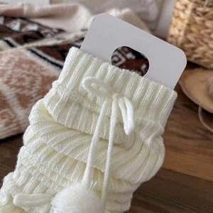 Close-up of cream knit leg warmers featuring vertical ribs, bow tie, and soft pom-poms