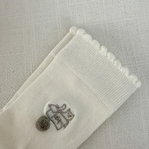Close-up of cream knit sock featuring cat embroidery, wavy edges and a grey 3D pom-pom detail