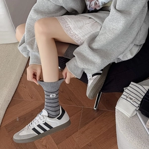 Dettaglio calzini grigi a righe con etichetta Bee e sneaker bianche su pavimento in legno