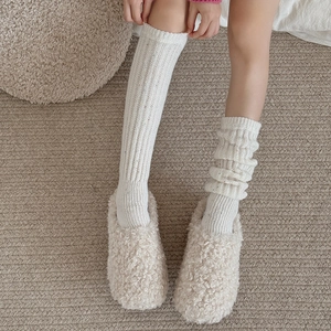 Cozy white ribbed knit leg warmers with plush cream slippers on a textured carpet