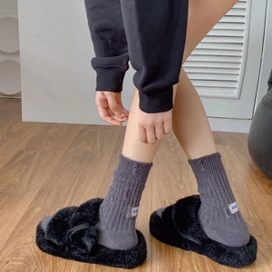 Close-up of black fuzzy cross-strap slides worn with grey socks on wooden floor