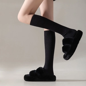 Woman wearing black ribbed knee-high cashmere socks with fuzzy plush slippers on grey background