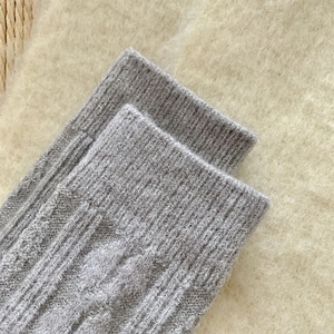 Close-up of light gray cable knit wool socks featuring ribbed cuffs on soft white carpet