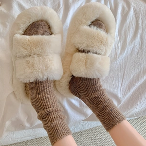 Close-up of fluffy beige faux fur slide slippers paired with brown knitted socks on a bed