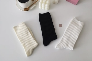Three pairs of ribbed crew socks in cream, black, and white on a minimalist desk setup