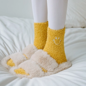 Close-up of yellow fleece socks with smiley embroidery and beige plush slippers on a bed