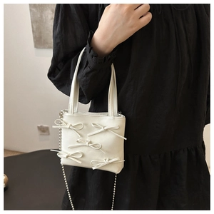 White mini bucket bag with 3D bows and chain strap held by a hand over black dress