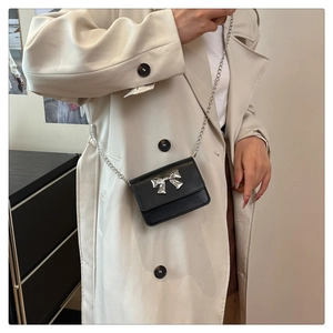 Close-up of black mini crossbody bag with silver bow clasp worn over a beige trench coat