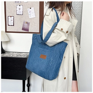 Detail of blue denim tote bag featuring a textured vertical pleat and brown leather logo patch
