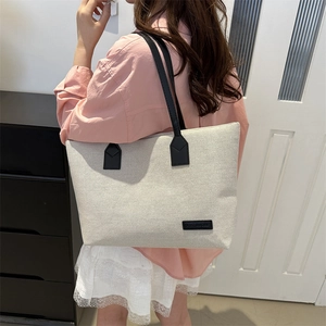 Large beige canvas tote bag with black shoulder straps and fashion metal logo plate