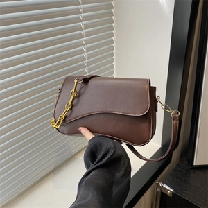 Brown minimalist baguette shoulder bag with gold chain and wavy flap in front of shutters