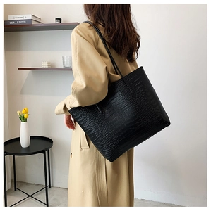 Model wearing black crocodile-embossed tote bag over a khaki trench coat in a cozy room