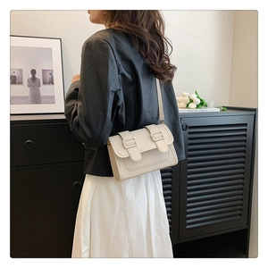 Detail of a creamy beige crossbody square bag with double buckles on a minimal indoor backdrop