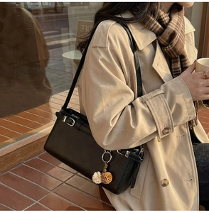 Detail of black rectangular leather shoulder bag with plush pendant on a beige trench coat