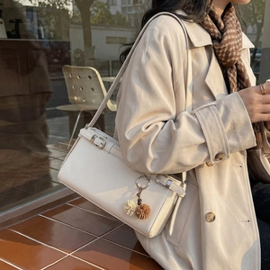Chic beige square crossbody bag with pompom charm shown on a stylish trench coat model