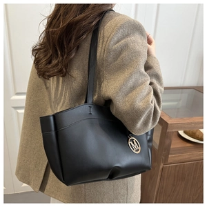 Detail of a large black faux leather tote bag with gold M-logo charm on a shoulder strap