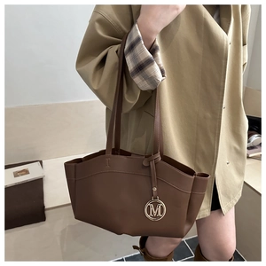 Detail of deep brown tote bag with metal M pendant and long shoulder strap worn by a person