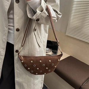 Brown suede half-moon crossbody bag with gold studs shown on model in urban setting