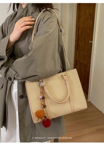 Close-up of beige woven tote bag with brown pom-pom charm and slender shoulder strap