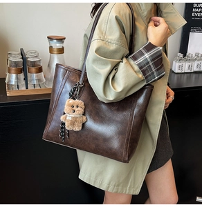 Brown oil wax leather tote bag with plush bear charm worn by model in a cafe setting