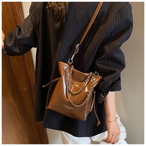 Brown faux leather bucket bag with chain detail and drawstring closure shown on shoulder