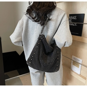 Detail of black denim tote bag featuring silver rivet trim and metal chain strap on model