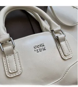 Détail du sac Boston blanc cassé montrant le cuir PU grainé et le logo en métal argenté