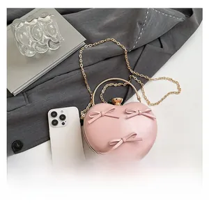 Pink heart-shaped clutch bag with bows and gold chain shown next to a smartphone