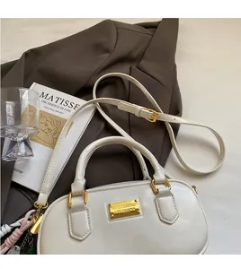 Detail der cremefarbenen Boston-Tasche mit Goldplakette auf braunem Blazer und Kunstbuch