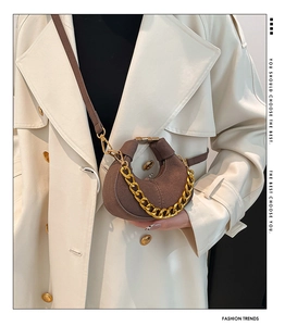 Detalle de bolso media luna en ante marrón con cadena dorada sobre gabardina blanca