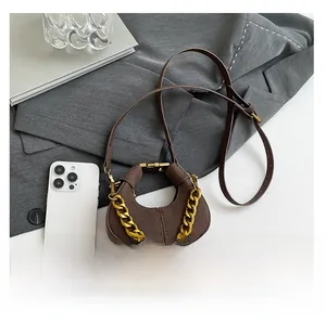 Mini bolso media luna de ante marrón con detalle de cadena dorada junto a un móvil