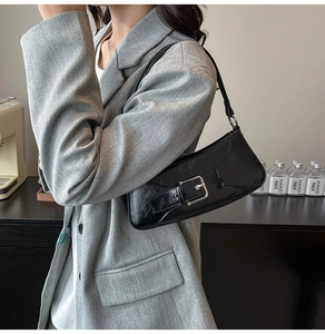 Close-up of a black glossy leather baguette bag with a silver buckle worn by a model