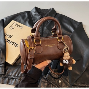 Detail of a brown cylindrical Boston bag with a plush monster charm and golden branding