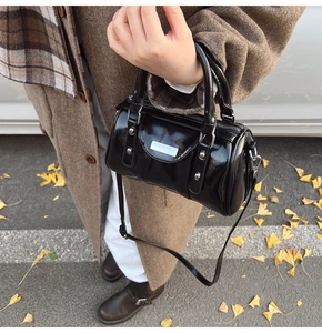 Detail of a shiny black barrel bag with silver hardware held by a model in autumn scenery