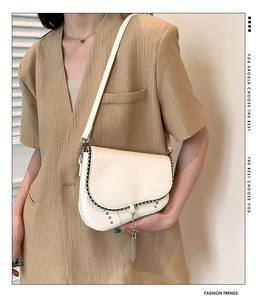 Détail d'un sac besace beige avec rivets argentés et pompon porté par un mannequin