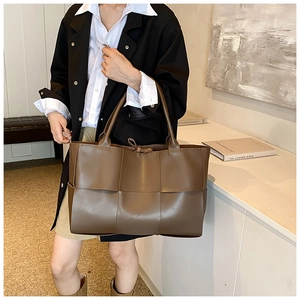 Model wearing a dark brown woven tote bag with wide straps in a minimalist studio setting