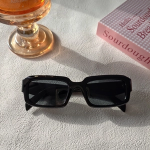 Détail de lunettes de soleil carrées en résine noire sur table blanche avec ombres
