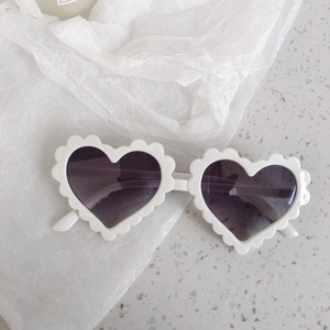 Detail view of white heart-shaped sunglasses with wavy scalloped frames on a textured base