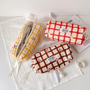 Three checkered cherry pencil pouches in yellow, red, and brown on a cozy desk setup