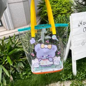Close-up of a clear PVC tote bag with cartoon bear print and yellow handles in a garden